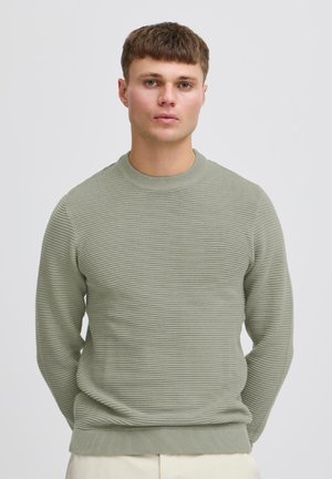 Jeune homme aux cheveux bruns courts portant un pull côtelé vert clair et un pantalon beige, debout devant un fond clair uni.