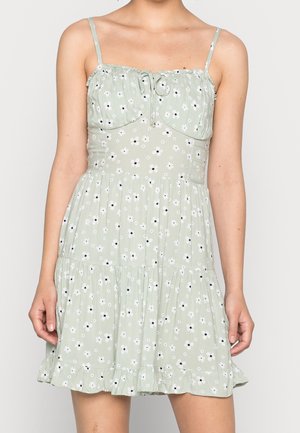 Robe sans manches vert clair avec fines bretelles, imprimé floral blanc, buste froncé avec lien, et ourlet à volants à plusieurs niveaux sous le milieu de la cuisse.