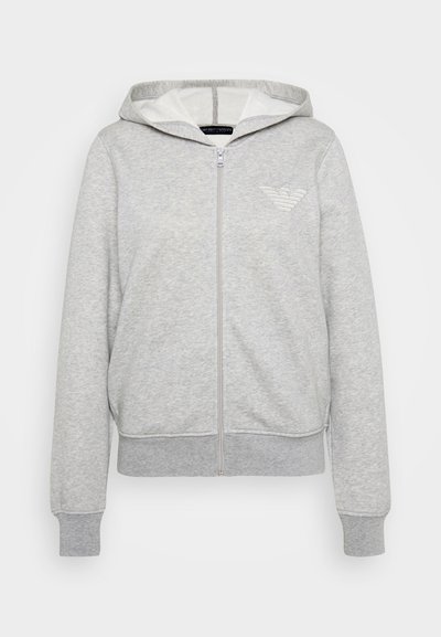 Gaiši pelēka zip-up kapuce, kas izgatavota no mīksta auduma, ar kapuci, priekšējām kabatām un izšūtu logotipu kreisajā krūtī.