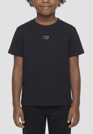 Dijete nosi jednobojnu crnu majicu kratkih rukava s malom kvadratnom značkom na prsima, stoji na svijetlosivoj pozadini.