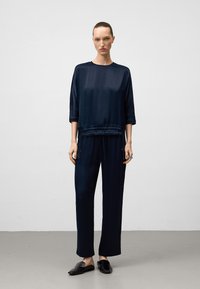 Kvinna som står inomhus iförd en navyblå satinblus med trekvartsärmar och matchande lösa byxor, med svarta tofflor.