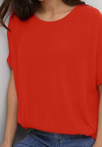 Blouse rouge à manches courtes en tissu léger et texturé. Encolure arrondie et coupe décontractée, associée à un jean en denim bleu.