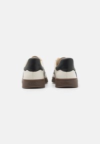 Lærersneakers i svart og kremfarge, med en strukturert brun gummisåle og med scallopdesign ved hælen.