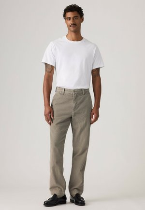 Mann mit kurzen lockigen Haaren und Schnurrbart, der ein weißes T-Shirt, beige Hose und schwarze Schuhe trägt, steht vor einem einfarbigen Hintergrund.