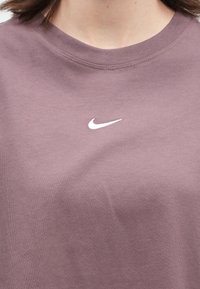 Κοντινό πλάνο ενός ατόμου που φοράει ένα μωβ κοντομάνικο μπλουζάκι με στρογγυλή λαιμόκοψη, με ένα μικρό λευκό λογότυπο Nike στο κέντρο του στήθους.