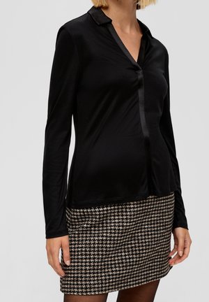 Femme portant un chemisier noir à manches longues et une jupe marron et noire à motif pied-de-poule sur un fond uni.