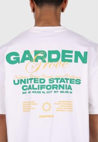Hvid bomulds T-shirt med stort grønt og gult grafisk tekst. Indeholder navne på Californiske byer og koordinater trykt på bagsiden.