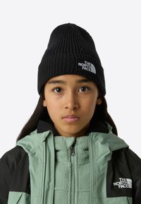 Svart strikket lue med logopatch, brukt med en grønn fleecefôret jakke med svart ytterstoff, synlig glidelås og hette.