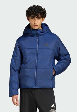 Junger Mann trägt eine blaue gesteppte Kapuzenjacke und schwarze Adidas-Hose, steht mit einer Hand in der Jackentasche.