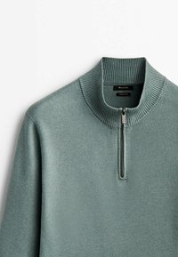 Pull-over en maille verte avec col montant côtelé et demi-fermeture éclair, montrant les détails du haut devant et du col sur fond blanc.