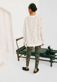 Felpa bianca con motivo decorativo, abbinata a leggings neri con motivi intricati e sneakers nere con suola bianca.