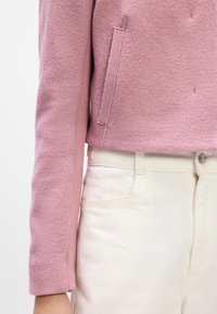 Jachetă din lână roz, cu un design scurt și o finisare texturată, asortată cu pantaloni din denim crem, care au buzunare frontale.