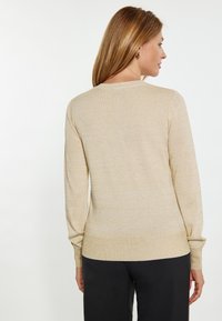 Beige gebreide trui met lange mouwen en een ronde halslijn, met een subtiele glans en een getailleerde taille, gecombineerd met zwarte broek.