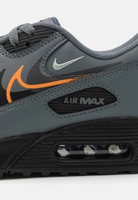 Grå sneakers med överdel i läder och orangea och svarta detaljer, snörning samt en synlig luftkuddsenhet i sålen.