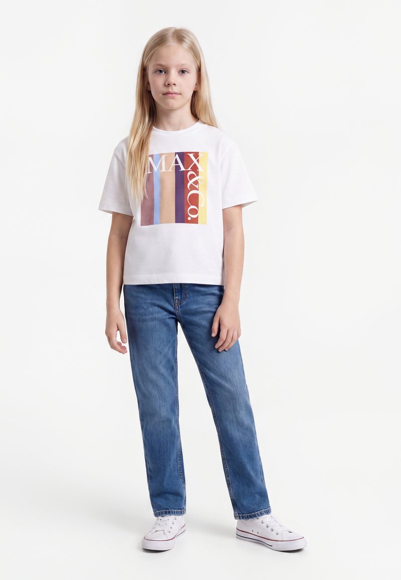 Jeune fille aux longs cheveux blonds portant un t-shirt blanc à motif, un jean bleu et des baskets blanches, debout devant un fond uni.