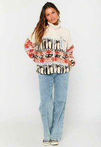 Fleece jacket in cream with multicolored palm tree pattern and blue accents. Full zip front, two pockets, relaxed fit. Light blue jeans.