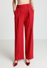 Rote, weit geschnittene Hose mit Plisseefalten, ausgestattet mit Seitentaschen und glatter Stofftextur. Kombiniert mit scharfen, spitz zulaufenden roten High Heels.