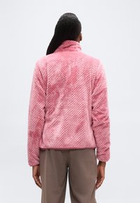 Άτομο με μακριά μαλλιά που φοράει μια υφασμάτινη ροζ φλις μπλούζα και καφέ παντελόνια, στέκεται γυρισμένο με την πλάτη προς ένα απλό φωτεινό φόντο.