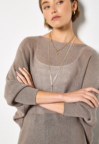 Leichter, taupefarbener, gestrickter Pullover mit lockerem Schnitt, weitem U-Boot-Ausschnitt und hochgekrempelten Ärmeln. Geschichtet mit goldenen Halsketten und subtiler Textur.