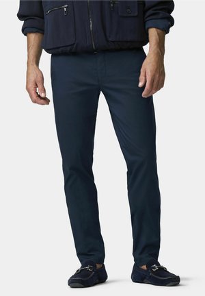 Mann trägt dunkelblaue, schmal geschnittene Hose, dunkelblaue Loafers und eine dunkelblaue Jacke mit Reißverschluss- und Knopftaschen, steht vor einem einfarbigen Hintergrund.