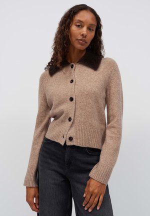 Donna con capelli ricci che indossa un cardigan beige con bottoni, colletto scuro e jeans neri su uno sfondo chiaro uniforme.