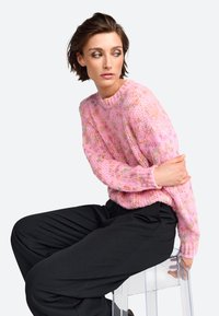 Pull en tricot rose avec une finition texturée, comportant des éclats multicolores. Modèle portant un pantalon large noir tout en étant assis sur une chaise transparente.