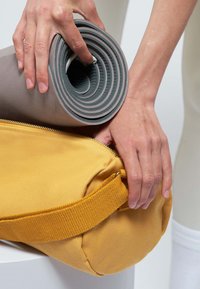 Bolso de deporte amarillo con un acabado texturizado, que cuenta con un zipper resistente y una correa tejida. Esterilla de yoga gris enrollada firmemente en la mano.