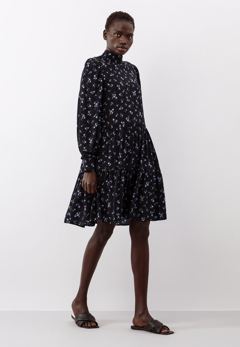 Vestido preto com um decote alto, mangas bufantes longas e uma saia em camadas. Apresenta pequenos padrões florais azuis por todo o vestido. Usado com sandálias pretas.