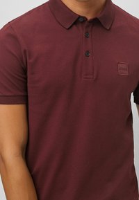 Burgundy polo skjorta i ett texturerat tyg med klassisk krage, tre svarta knappar och en liten präglad logotyp på bröstfickan.