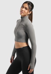 Gekürzte graue Sportjacke mit hohem Kragen und langen Ärmeln, die eine glatte Textur und eine taillierte Passform aufweist. Kombiniert mit schwarzen Leggings.