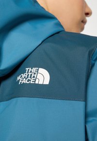 Blue hooded jacket with a smooth texture. Features a dark blue yoke and a white logo on the back. Stitched detailing is visible.