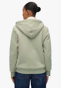 Vrouw met een bun, gekleed in een lichtgroene hoodie en blauwe jeans, gezien van achteren tegen een witte achtergrond.