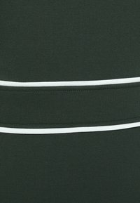 Tissu vert foncé en gros plan avec deux bandes blanches parallèles et des coutures visibles entre les bandes.