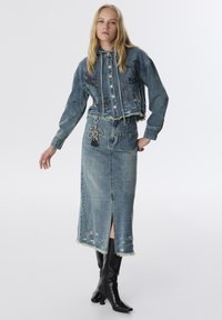 Jeansjacke mit fransigen Kanten, bestickten Details und Knöpfen, kombiniert mit einem langen, zerstörten Jeansrock mit Schlitz vorne. Schwarze knielange Stiefel.