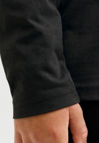 Fekete, hosszú ujjú ing, puha tapintással. Közelkép a mandzsettáról, amelyen látszik a varrott szél és a sima felület, egy kéz fogja a szövetet.