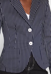 Blazer rayé navy avec des lignes blanches verticales, deux boutons blancs, revers crantés et deux poches avant. Convient pour un vêtement formel ou professionnel.
