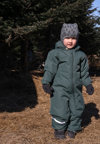 Mono de nieve de color verde oscuro con cremallera frontal, puños elásticos y ribete blanco. Acompañado de un gorro gris con estampado y guantes negros.