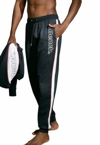 Schwarze Jogginghose mit weißen und kastanienbraunen Seitenstreifen, ausgestattet mit einem Kordelzugbund und bedrucktem Logo. Das Model hält eine passende Jacke.