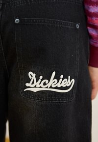 Pantalons en denim noir avec un logo "Dickies" brodé en blanc sur la poche arrière, au toucher lisse et à la coupe classique.