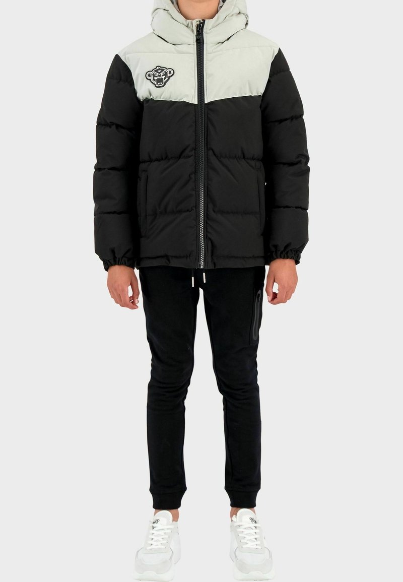 Enfant portant une doudoune à capuche noire et beige, un pantalon de jogging noir et des baskets blanches, debout devant un fond blanc.