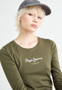 Maslinasto zelena majica dugih rukava s okruglim izrezom i bijelim logotipom "Pepe Jeans London". Model nosi sivi šešir i ima kratku, valovitu kosu.