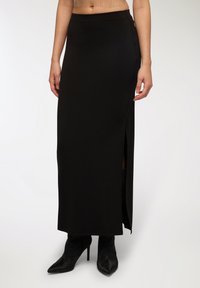 Gonna nera maxi realizzata in tessuto liscio, con uno spacco laterale e una vita aderente. Indossata con tacchi a punta neri.