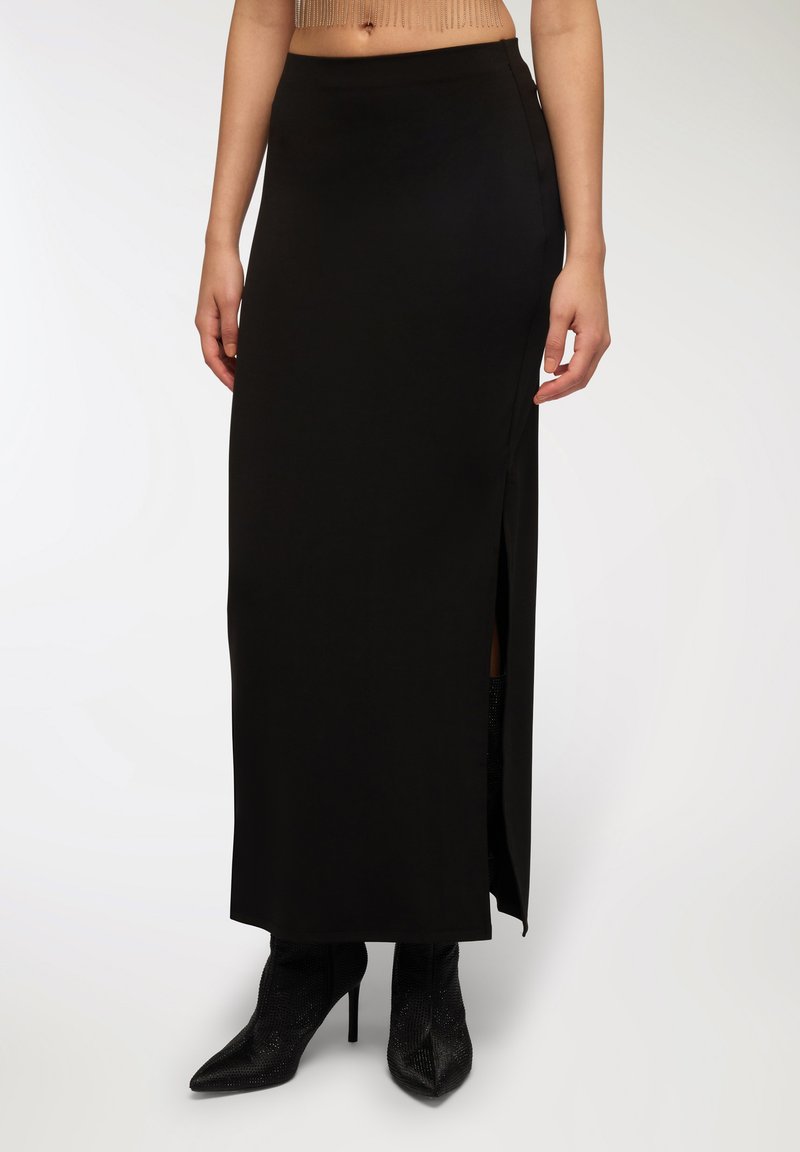 Gonna nera maxi realizzata in tessuto liscio, con uno spacco laterale e una vita aderente. Indossata con tacchi a punta neri.