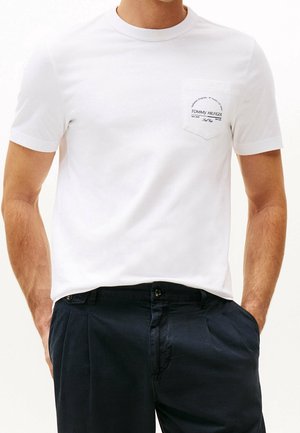 Mężczyzna ubrany w białą koszulkę z krótkim rękawem z małą kieszonką i czarnym napisem po lewej stronie klatki piersiowej, w ciemnoniebieskich spodniach, z rękami w kieszeniach.