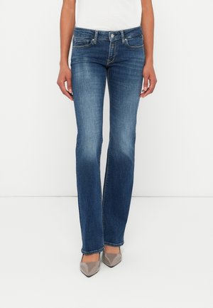 Femme portant un jean bootcut taille mi-haute bleu foncé avec des détails délavés et des talons hauts taupe pointus, debout sur fond blanc.