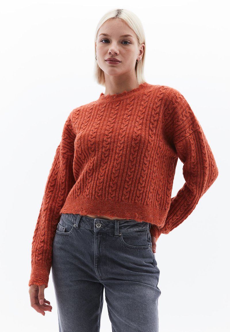 Gehaakte oranje sweater met textuur kabelpatroon, ronde halslijn en cropped lengte; gedragen met grijze high-waisted jeans.