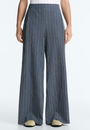 Osoba ubrana w luźne, granatowe spodnie z wysokim stanem, ozdobione cienkimi, pionowymi białymi paskami, oraz beżowe buty na jednolitym tle.