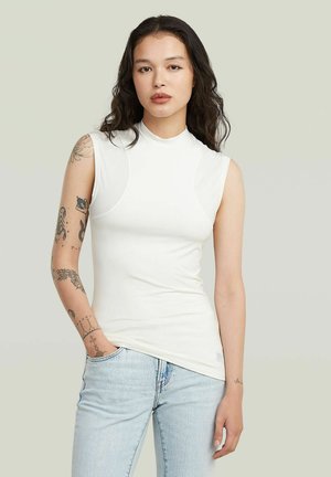 Giovane donna con lunghi capelli scuri che indossa un top bianco senza maniche e jeans azzurri, con tatuaggi visibili sul braccio sinistro.