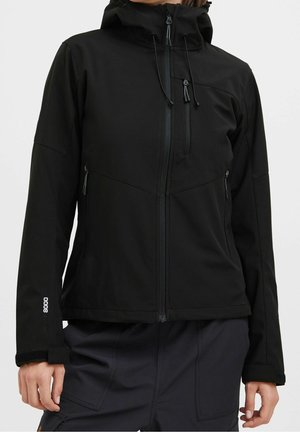 Persoon draagt een zwarte waterdichte jas met capuchon, ritsen aan de voorkant en op de borst, en verstelbare manchetten, gecombineerd met een donkere broek.