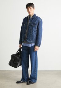 Jeansjacka med två bröstfickor, blå vida jeans och svarta läderskor. Åtföljd av en svart väska, bakgrunden är enfärgad.